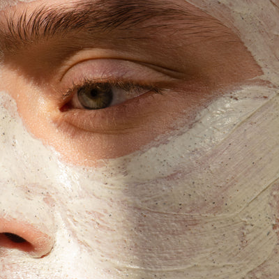 Hand squeezing blue Double Down Face cleanser and Exfoliating Mask tube and close up of man's face with mask