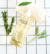 Botanical Protein Complex Shampoo bottle with gold liquid on white tile surface with rosemary and sage sprigs and loofah