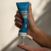 Double Down Face Cleanser and Exfoliating Mask blue tube in man's hand. One hand squeezing tube and gray cream into other hand